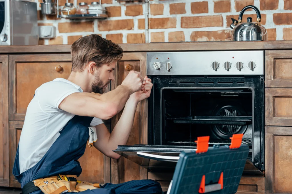 Male technician kneels by an open oven, hands on the door, inspecting the interior in a rustic kitchen with a brick backsplash and tool belt nearby