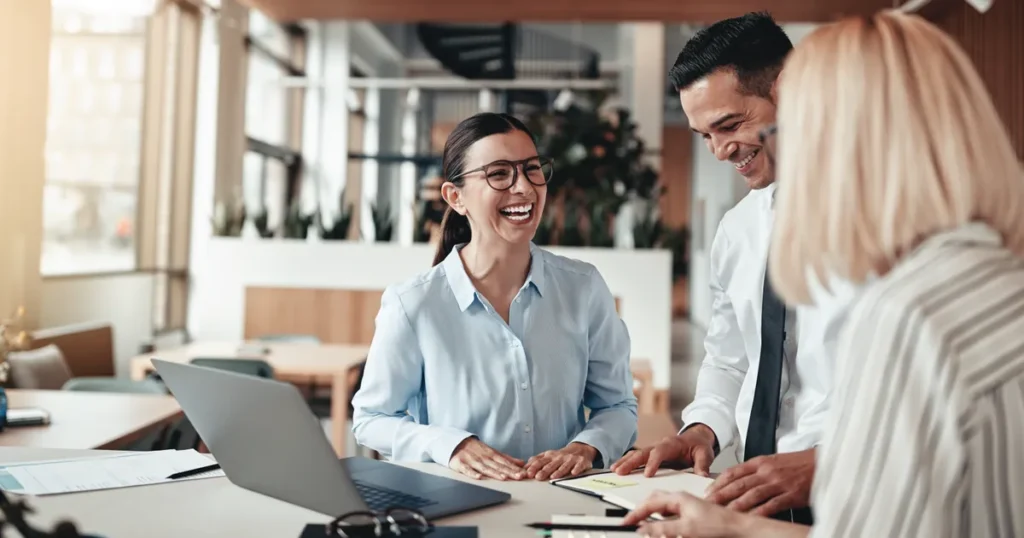 Three coworkers in a bright office laugh and review documents at a shared desk with a laptop nearby.