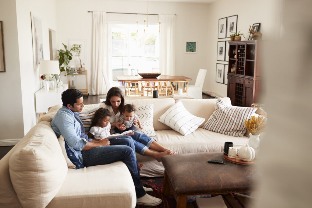Family cuddled on a light sofa reading a book together in a bright living room. Two children with their parents outdoorsy light, cozy home.