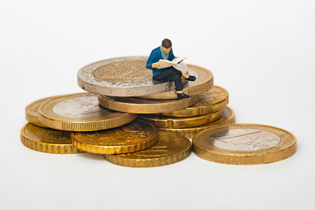 Miniature figure in a blue jacket sitting on stacked coins and reading a newspaper to illustrate finance concepts.