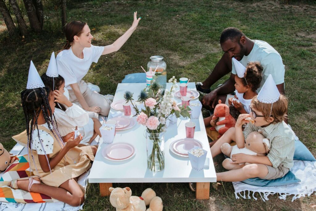 Group of adults and children having a picnic on blankets around a low white table with pastel pink dishes and cups on a sunlit lawn.