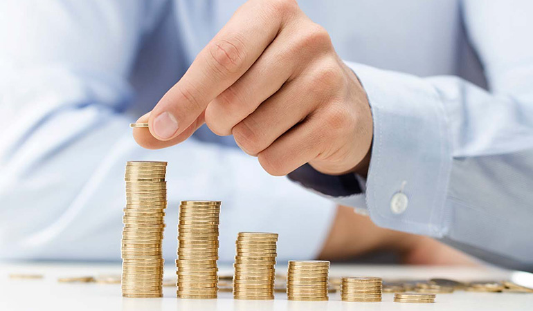Person places a coin on a tall stack, illustrating saving and financial growth across several coin towers on a table.