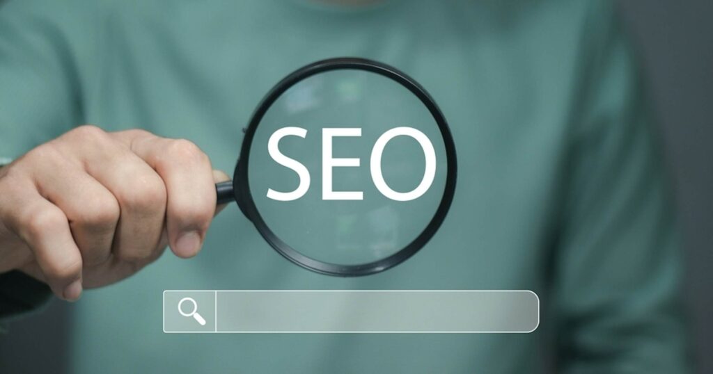 Magnifying glass held by a hand focusing on the word SEO, with a faint search bar beneath (SEO concept)
