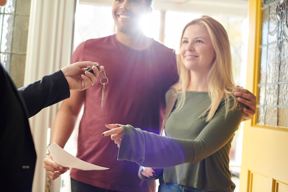 Couple receives house keys from a real estate agent at their new home, smiling by the front door.