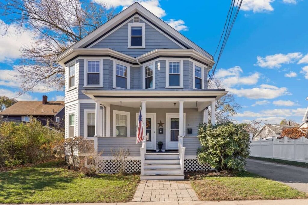 Front view of a blue two-story house with a sunny porch, white columns, and an American flag by the entrance.